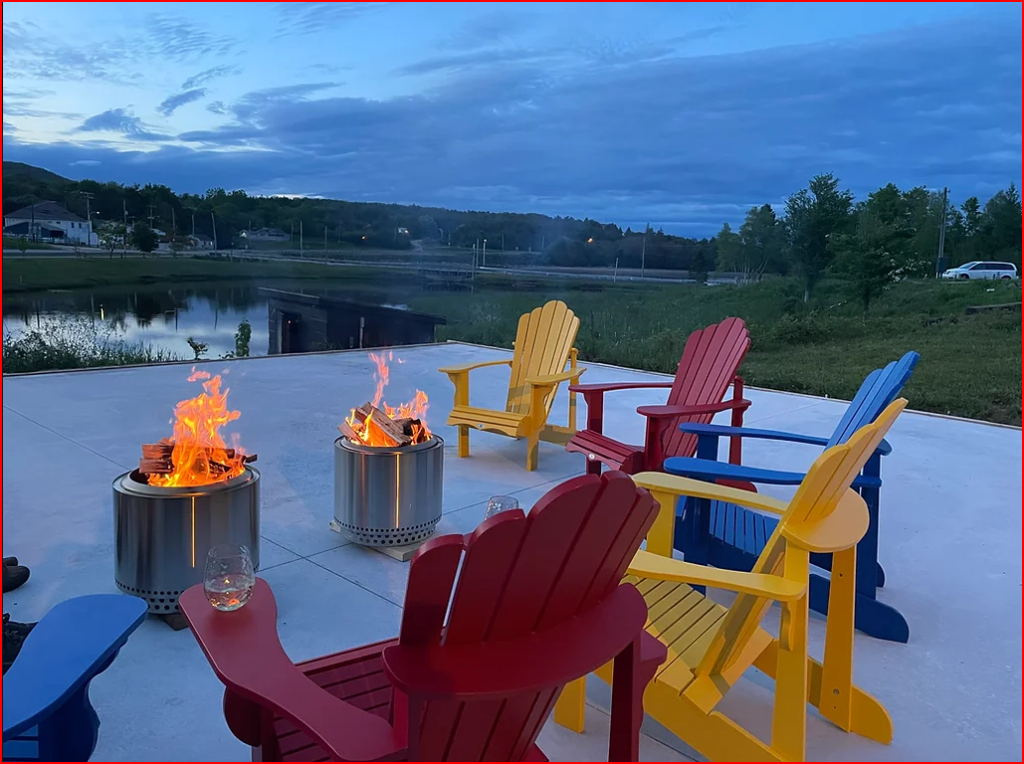 Six colorful Adirondack chairs surround two fire pits on a patio near a pond at dusk, with drinks on the chairs and a scenic landscape in the background.