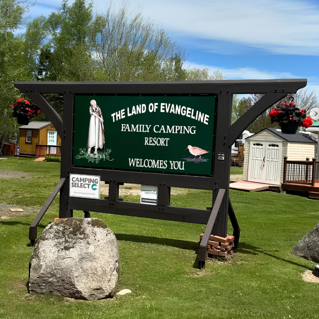 A large green sign reads “The Land of Evangeline Family Camping Resort Welcomes You,” with images of a woman and a bird, on a grassy area with buildings and trees in the background.