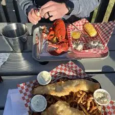 A person prepares to eat a cooked lobster with corn and potatoes, while a tray of fried fish, curly fries, and tartar sauce sits in the foreground.