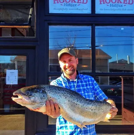 A man in a blue plaid shirt and cap stands outside a shop holding a large fish with both hands, smiling at the camera.