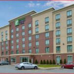 A five-story Holiday Inn Express & Suites hotel with a red and beige exterior, parked cars in front, and a mostly clear sky overhead.