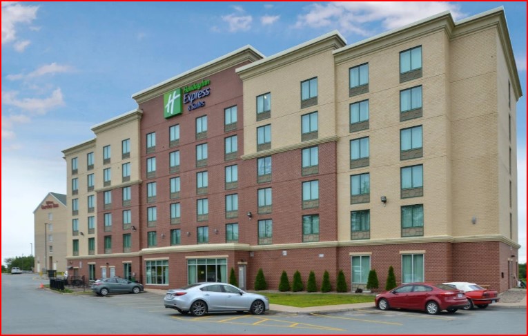 A five-story Holiday Inn Express & Suites hotel with a red and beige exterior, parked cars in front, and a mostly clear sky overhead.