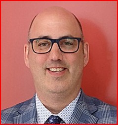 A man with glasses, wearing a checked suit jacket, white shirt, and tie, poses in front of a plain red background.
