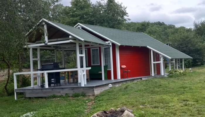 A small red and white cabin with a green roof sits on grassy land, featuring a covered porch and a stone fire pit in front, surrounded by trees under a cloudy sky.