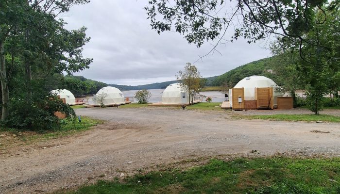 Four white geodesic domes are set on a gravel lot near a lake, surrounded by trees and hills under a cloudy sky.