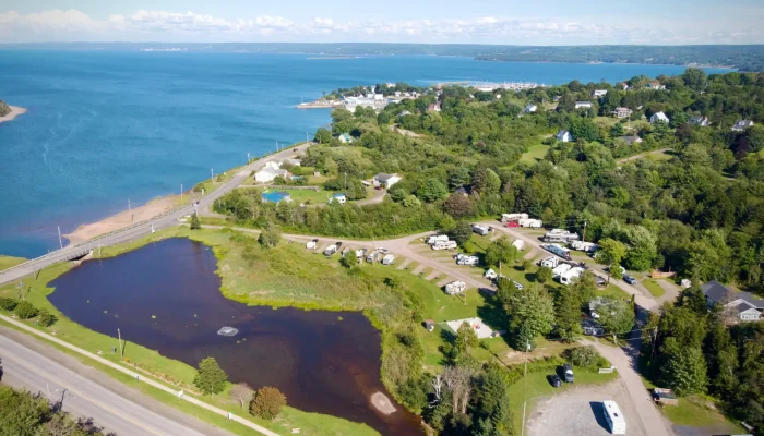 Aerial view of a coastal campground with RVs and a pond near a road, bordered by green trees and overlooking a bay with a distant shoreline.