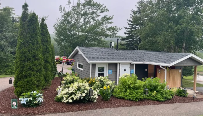 A small public restroom building is surrounded by landscaped flower beds, tall shrubs, and trees, with a paved path leading past it on a cloudy day.