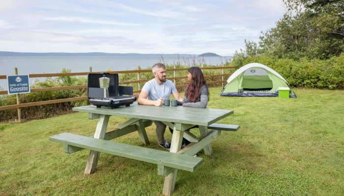 A man and a woman sit at a green picnic table with coffee mugs and a portable stove, near a tent and overlooking a body of water at a campsite.