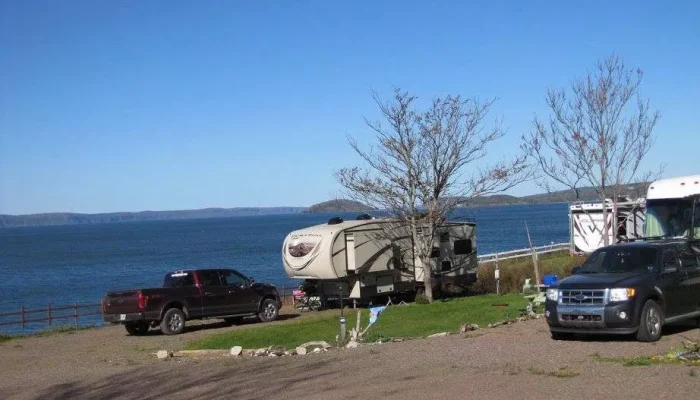 A camper, pickup truck, and SUV are parked near leafless trees beside a large body of water under a clear blue sky.