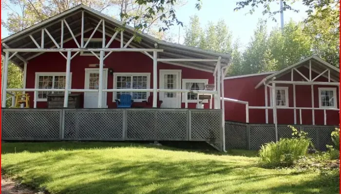 Red wooden house with white trim and a covered porch, surrounded by green grass and trees on a sunny day.