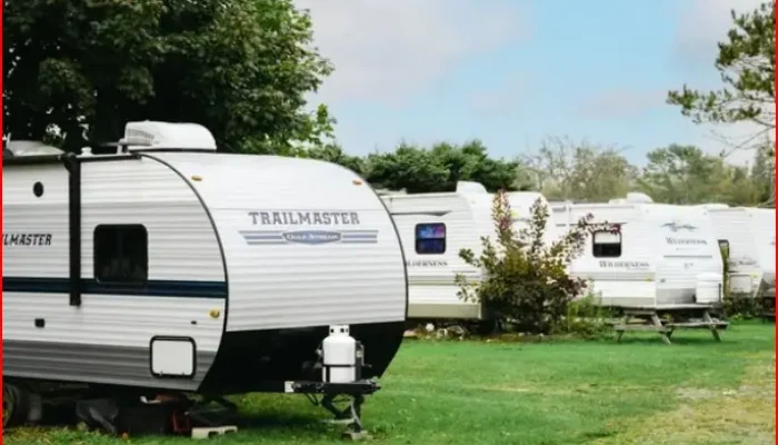 Several white travel trailers are parked on grassy lots with picnic tables nearby, surrounded by trees under a blue sky.