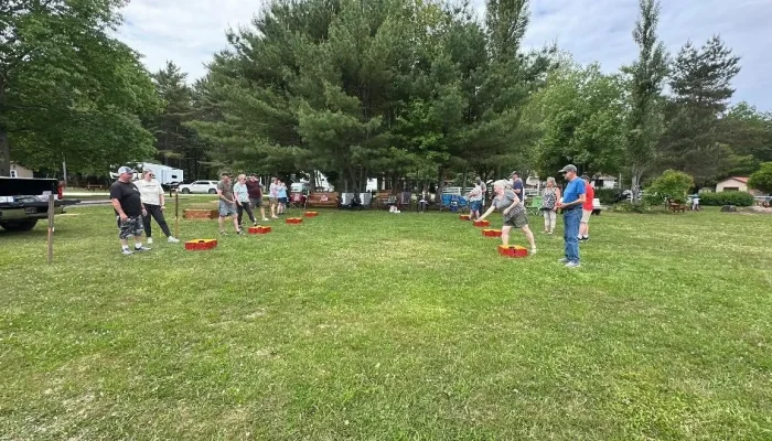 A group of people are playing yard games on a grassy field surrounded by trees, with some RVs and picnic tables in the background.