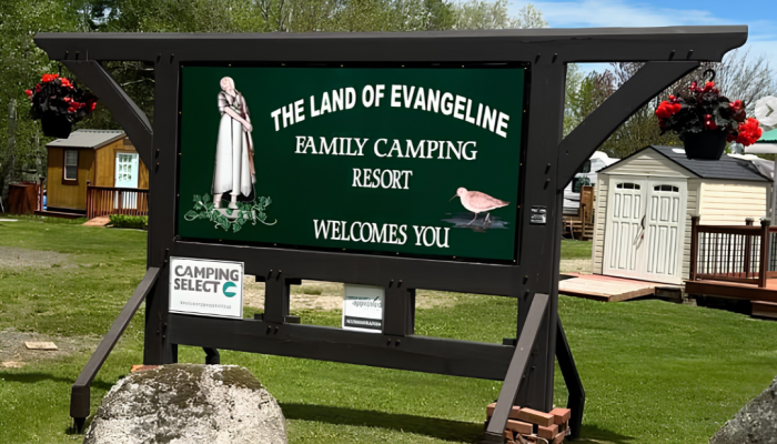 A large green sign reads “The Land of Evangeline Family Camping Resort Welcomes You,” with images of a woman and a bird, on a grassy area with buildings and trees in the background.