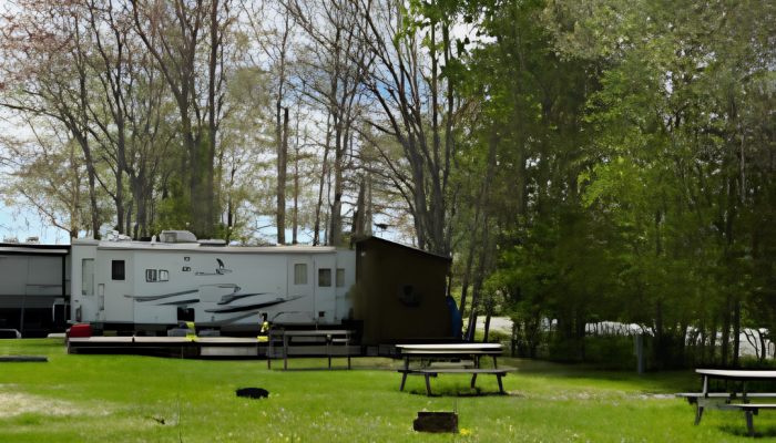 A white RV is parked on a grassy area near trees, with picnic tables and a fire pit in the foreground.