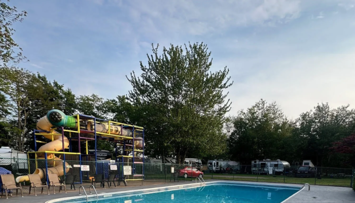 Outdoor swimming pool with metal ladders, surrounded by trees and RVs. A playground with yellow slides is visible in the background under a partly cloudy sky.