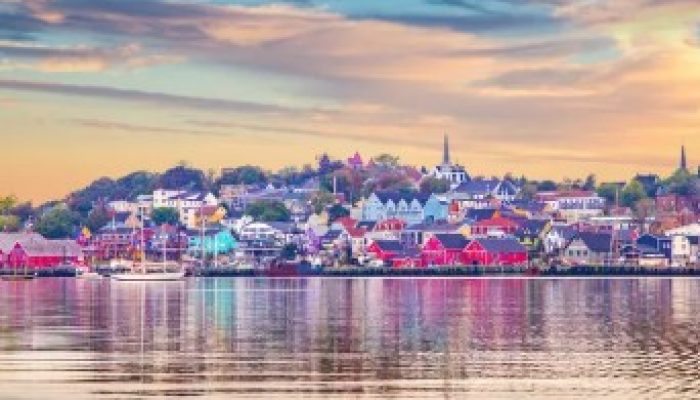 A colorful waterfront town with houses and buildings reflected in calm water under a cloudy, pastel sky at sunset.