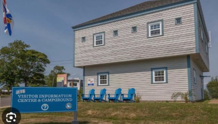 A two-story gray and blue building with a sign in front reading "Visitor Information Centre & Campground" and several blue chairs on the lawn.