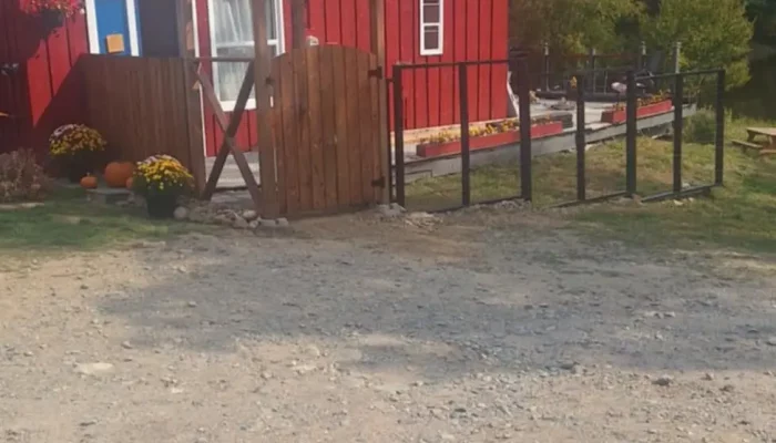 A red and blue building with an "Office" sign, a wooden gate, potted flowers, and a gravel driveway; trees and hills are visible in the background.