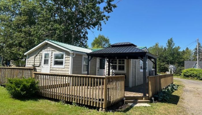 Single-story cottage with a green metal roof, wooden deck, and a black gazebo in a grassy yard on a sunny day.