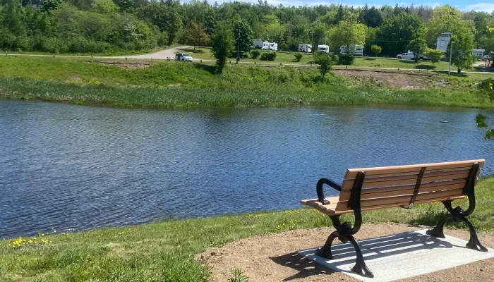 A wooden bench faces a calm river, with grassy banks and trees in the background. Several RVs are parked on the opposite side near a road under a blue sky.