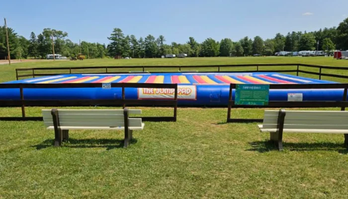 Two benches face a fenced-in, colorful inflatable jump pad called "The Jump Pad" on a grassy field with trees and a clear sky in the background.
