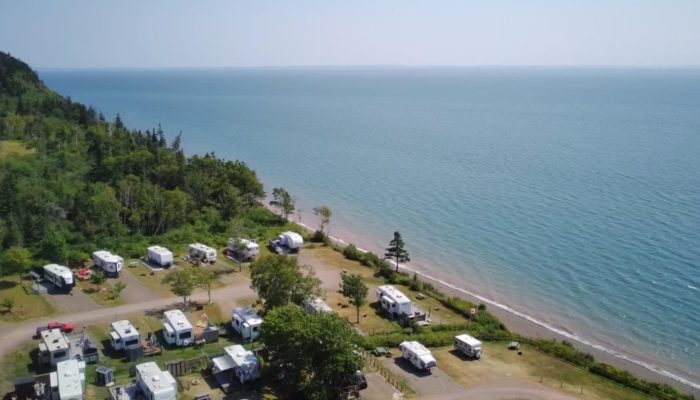 Aerial view of a lakeside RV campground with several white campers parked near the water and surrounded by trees. The shoreline curves along a calm, blue lake under a clear sky.