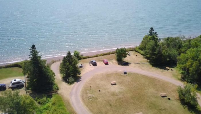 Aerial view of a lakeside parking area with a few cars, benches, and trees, bordered by a curved dirt road and overlooking a large body of water.