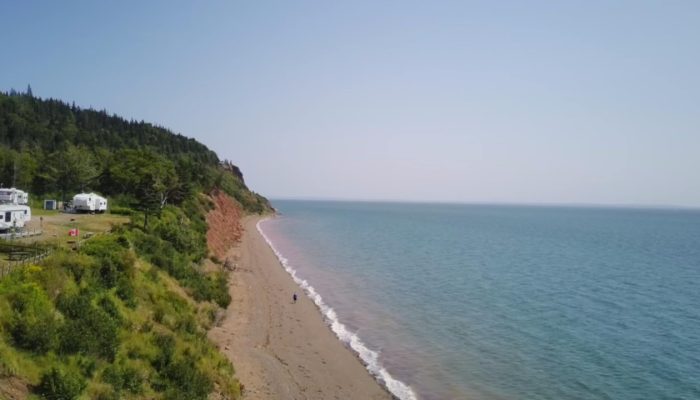 A sandy beach with gentle waves borders a forested hillside with a few RVs and trailers parked near the edge, under a clear blue sky.