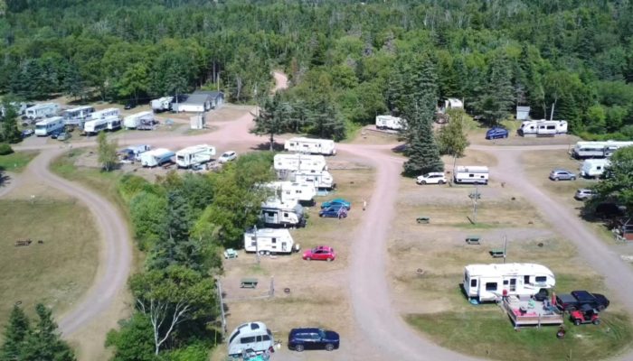 Aerial view of an RV park with several parked trailers and vehicles, surrounded by trees and winding dirt roads on a sunny day.