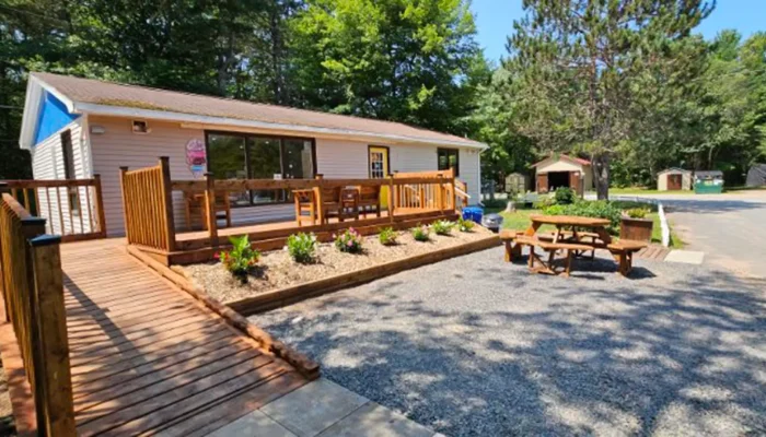 A single-story building with a wooden ramp and porch, flower beds, a picnic table, and surrounding trees on a sunny day.