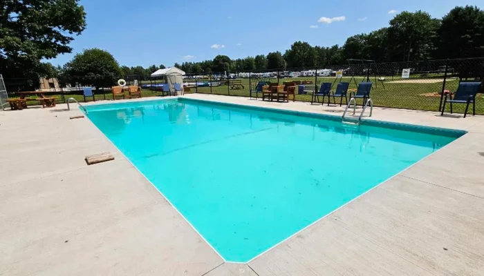 Outdoor swimming pool with clear water, surrounded by a concrete deck, lounge chairs, tables, and a black chain-link fence; trees and blue sky in the background.